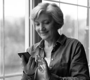 Middle-aged woman smiling at smartphone by window in serene black-and-white setting.