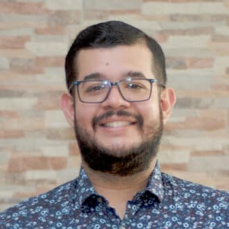 Portrait of juan camacho smiling, with glasses and floral shirt against a brick wall backdrop.