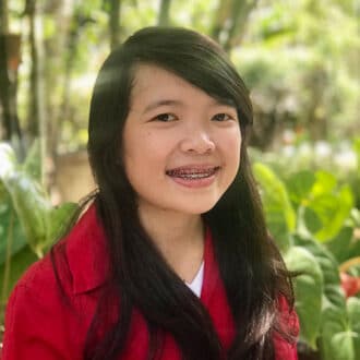 Asian woman in red jacket smiling outdoors, wearing braces, with a natural green backdrop.