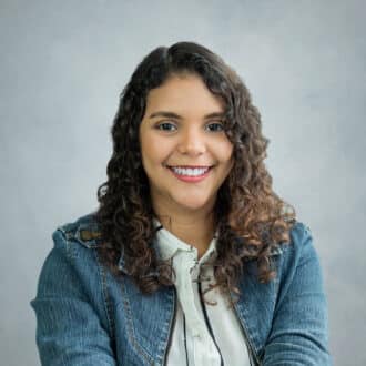 Professional headshot of confident woman with curly hair, smiling in denim jacket, light background.