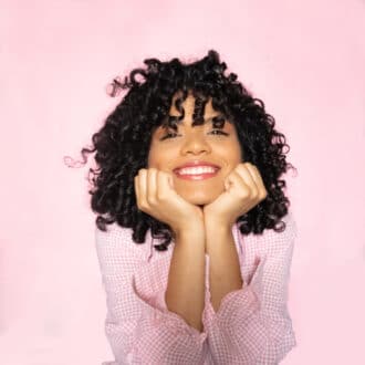 Joyful curly-haired person smiling broadly in polka-dot top against pink background.