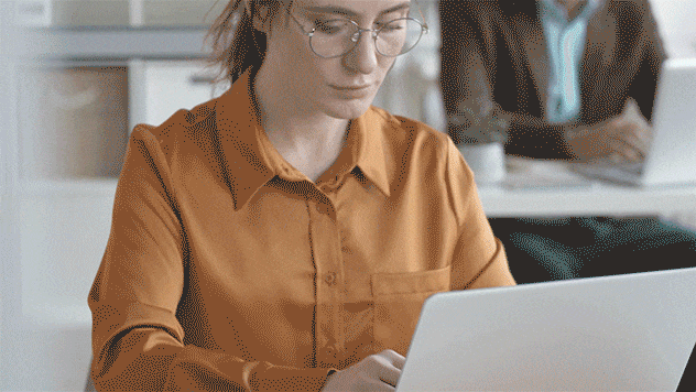 Professional in yellow shirt works intently on laptop in a bright, modern office setting.